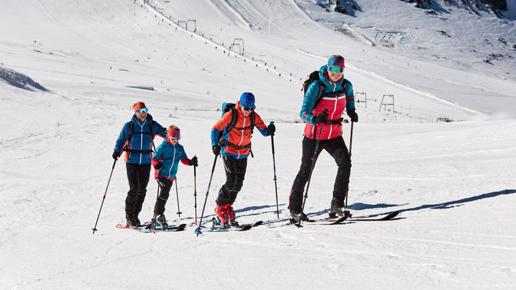 Vier Personen steigen mit Skiern und Skistöcken einen verschneiten Berghang hinauf. Sie tragen Winterkleidung mit Jacken, Hosen, Handschuhen und Helmen. Im Hintergrund sind schneebedeckte Hänge und Skiliftanlagen zu sehen