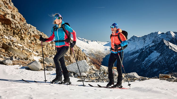 Zwei Skifahrer steigen einen schneebedeckten Berg hinauf. Sie tragen Winterkleidung, Rucksäcke und schwarze Hosen. Die linke Person hat eine rote Jacke mit blauen Ärmeln, die rechte eine orangefarbene Jacke mit blauen Ärmeln. Im Hintergrund sind verschneite Berge sichtbar