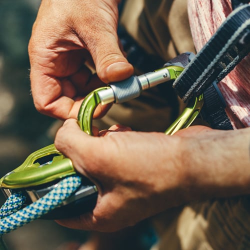 Nahaufnahme von Händen, die einen grünen Karabiner sichern. Der Karabiner ist mit einem blau-weiß gemusterten Kletterseil und einem schwarz-grauen Gurt verbunden.