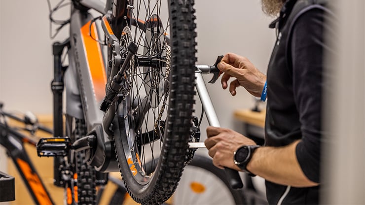 HERVIS Bikeservice Eine Person arbeitet an einem beigefarbenen Fahrrad in einer Fahrradwerkstatt oder einem Reparaturbereich. Das Gesicht der Person ist unscharf, und der Fokus liegt auf dem Hinterrad und dem Getriebemechanismus des Fahrrads. Im Hintergrund sind weitere Fahrräder und Ausrüstungen zu sehen.