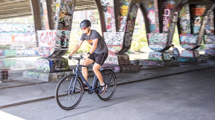 Person fährt mit dem Fahrrad unter einer Brücke, deren Pfeiler und Wände mit bunter Graffiti-Kunst bedeckt sind; die Person trägt Helm, T-Shirt, Shorts und Sneakers.