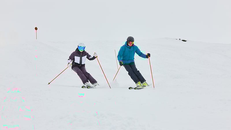 Zwei Skifahrer:innen fahren parallel die Piste hinunter und bewegen sich synchron.