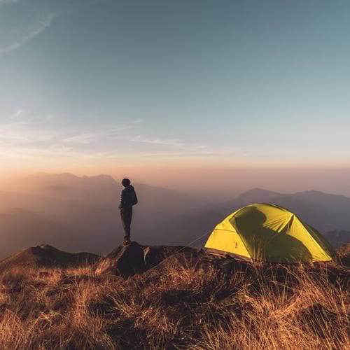 Person steht auf felsigem, mit trockenem Gras bewachsenem Gelände und blickt bei Sonnenauf- oder -untergang auf eine Berglandschaft. In der Nähe ist ein gelbes Zelt aufgebaut. Der Himmel ist klar mit wenigen Wolken – die Szene wirkt ruhig und abenteuerlich.