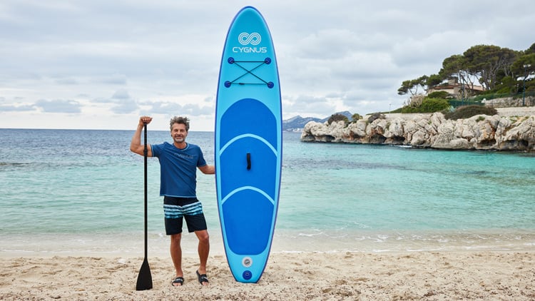 Eine Person steht am Sandstrand neben einem blauen Paddleboard der Marke CYGNUS und hält ein Paddel. Im Hintergrund sind türkisfarbenes Wasser, Felsen, Bäume und ein bewölkter Himmel zu sehen.