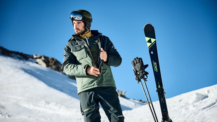 Skijacke Person in Wintersportausrüstung mit Helm und Skibrille steht auf einem verschneiten Hang, neben ihr stehen aufrecht Skier und Stöcke im Schnee. Im Hintergrund ist ein klarer blauer Himmel und schneebedecktes Gelände zu sehen