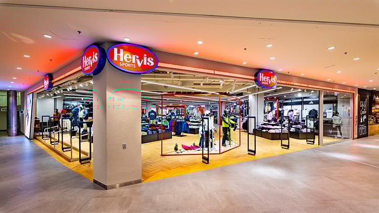 Die Fassade des Hervis Sports Stores Graz Cityparl. Der Store hat ein modernes Design mit großen Glasfenstern und -türen, die Einblick in das Innere gewähren. Im Inneren sind verschiedene Sportgeräte und Kleidungsstücke auf Regalen und Ständern ausgestellt. In der Nähe des Eingangs stehen Schaufensterpuppen, die Sportbekleidung tragen. Der Name 'Hervis Sports' ist auf beleuchteten Schildern über dem Eingang gut sichtbar.