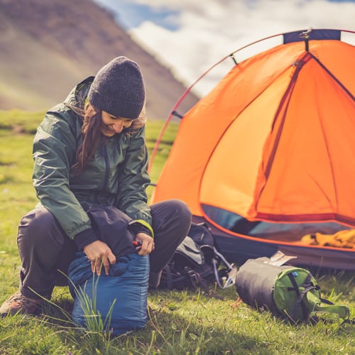 Person in Outdoor-Kleidung mit Mütze und grüner Jacke sitzt auf einer Wiese und packt eine blaue Tasche. Im Hintergrund steht ein orangefarbenes Zelt, dahinter Berge und ein teils bewölkter Himmel. Das Gesicht der Person ist nicht erkennbar