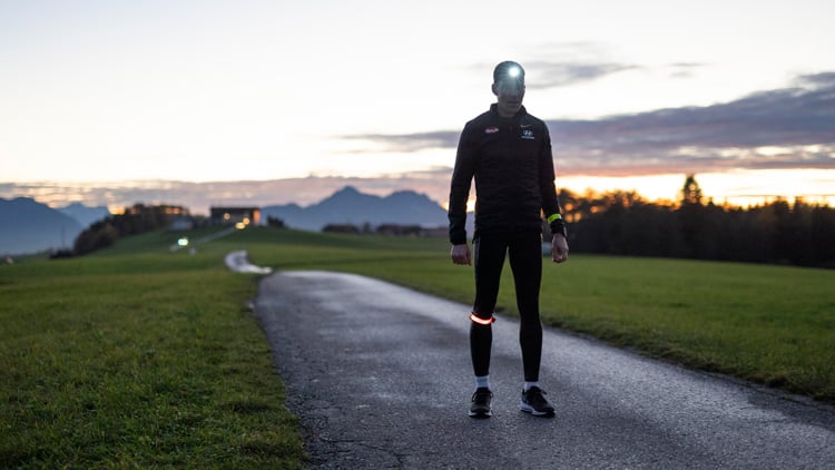 Person in dunkler Sportkleidung mit Stirnlampe und reflektierenden Bändern steht auf einem gepflasterten Weg in einem offenen Feld bei Dämmerung; im Hintergrund sind Berge und Bäume vor einem wolkigen Himmel zu sehen