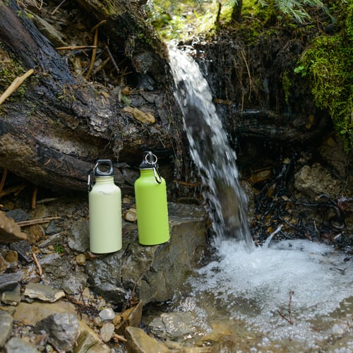 Zwei grüne Wasserflaschen mit Metallclips stehen auf einem Felsen nahe einem kleinen Wasserfall in einem moosbedeckten Waldgebiet