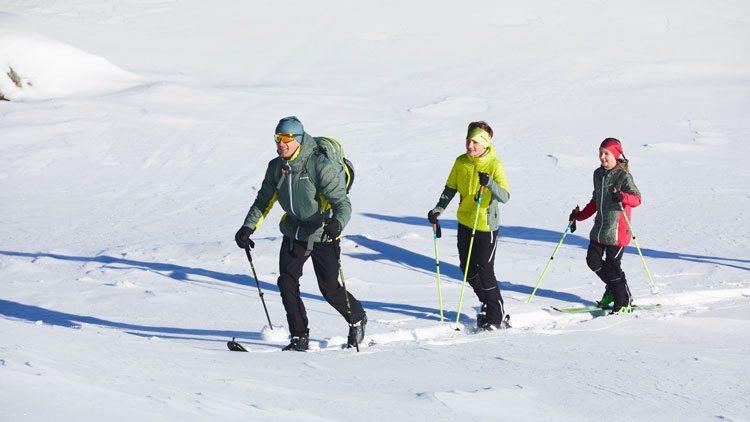 Drei Personen steigen mit Skiern einen verschneiten Hang hinauf. Sie tragen Winterkleidung mit Jacken, Hosen, Handschuhen und Mützen. Die erste Person trägt eine blaue Jacke und Mütze mit schwarzer Hose, die zweite eine gelbe Jacke mit schwarzer Hose und die dritte eine rote Jacke mit schwarzer Hose. Alle nutzen Skistöcke zur Unterstützung. Die Landschaft ist schneebedeckt und der Himmel klar.