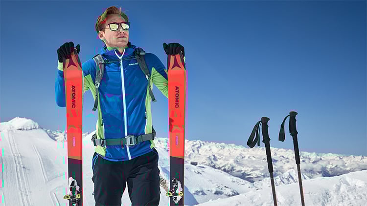Person steht auf einem verschneiten Berg und hält zwei rot-schwarze Skier der Marke ‚ATOMIC‘. Sie trägt eine blau-grüne Jacke, schwarze Handschuhe und einen Rucksack. Rechts im Schnee stecken zwei Skistöcke. Im Hintergrund sind schneebedeckte Berge unter klarem blauem Himmel zu sehen