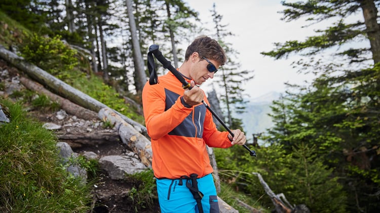 Person mit orangefarbener Jacke und blauer Shorts steht mit Wanderstock in einem bewaldeten Gebiet. Der Hintergrund zeigt Bäume und umgestürzte Baumstämme, was auf einen Wanderweg hinweist. Das Gesicht der Person ist unscharf