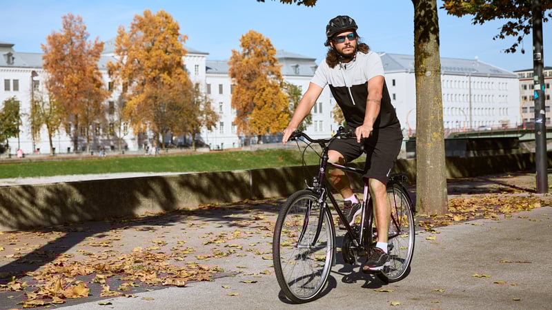 Person mit Helm, Sonnenbrille und Radkleidung fährt auf einem asphaltierten Weg mit einem Citybike. Im Hintergrund herbstlich gefärbte Bäume und Gebäude, auf dem Boden liegen verstreute Blätter.