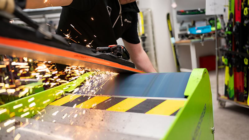 Auf dem Bild ist zu sehen, wie ein Ski in einer Werkstatt maschinell geschliffen wird. Dabei sprühen Funken, während die Kanten des Skis über die Schleifmaschine gezogen werden.