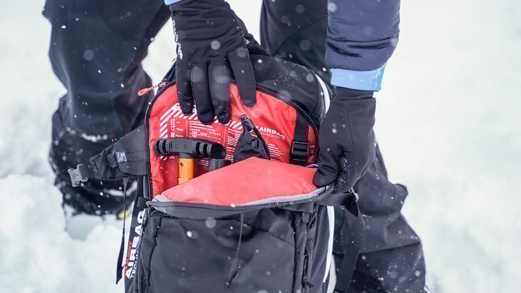 Person in Winterkleidung und schwarzen Handschuhen öffnet einen schwarzen Rucksack in verschneiter Umgebung. Im Rucksack befinden sich ein orangefarbenes Werkzeug und ein Lawinenairbagsystem mit rotem Stoff