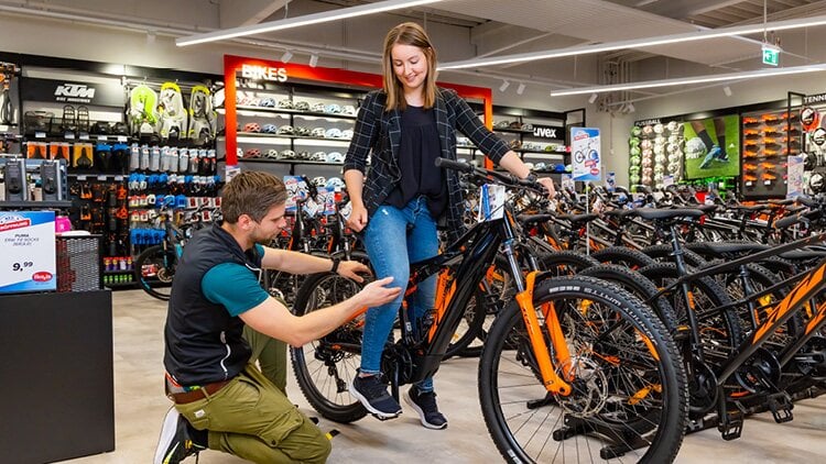 Eine Person in einem Fahrradgeschäft wird von einem Mitarbeiter unterstützt. Die Person sitzt auf einem schwarz-orangenen Fahrrad, während der Mitarbeiter kniet und das Fahrrad einstellt. Im Hintergrund sind verschiedene Fahrradzubehörteile, Helme und andere Fahrräder zu sehen.