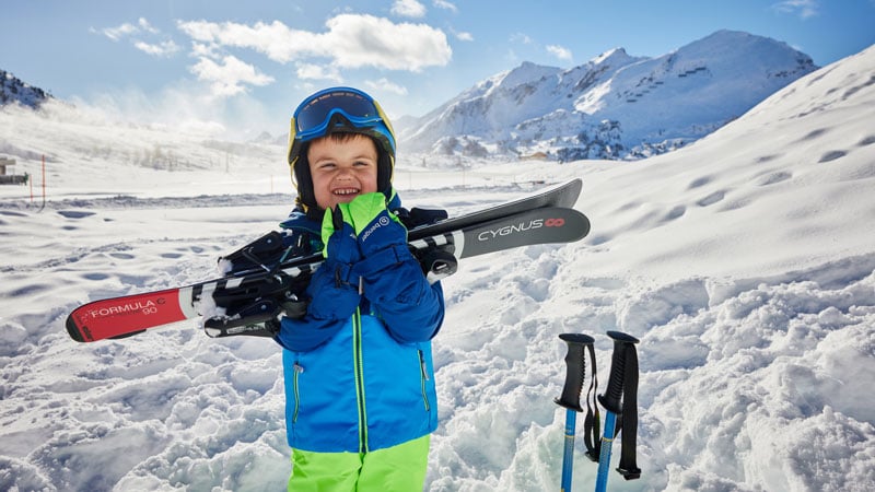 Kind in Winterkleidung mit blauer Jacke und grüner Hose steht in einem verschneiten Gebirgsgebiet und hält zwei Skier – mit der Aufschrift ‚FORMULA 90‘ und  ‚CYGNUS‘. Rechts im Schnee stecken zwei Skistöcke
