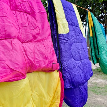 Mehrere bunte Schlafsäcke in Farben wie Pink, Gelb, Lila, Grün und Blau hängen draußen an einer Wäscheleine. Im Hintergrund sind Gras und Bäume zu sehen