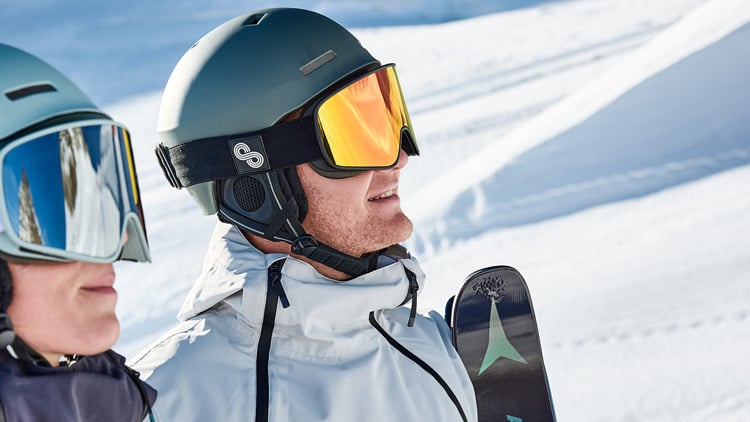 Zwei Personen in Winter-Sportkleidung mit Helmen und Skibrillen stehen auf einer verschneiten Piste. Eine Person hält Ski. Im Hintergrund sind Schneespuren zu sehen
