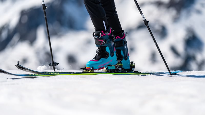Eine Person steht mit Tourenskischuhen und Tourenski im Schnee und stützt sich auf Skistöcke.