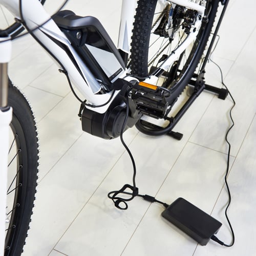 Weißes E-Bike mit schwarzen Akzenten steht auf einem Ständer und ist über ein Kabel mit einem Ladegerät verbunden; das Ladegerät liegt auf dem Boden, der Stecker ist im Motorbereich des Fahrrads eingesteckt.