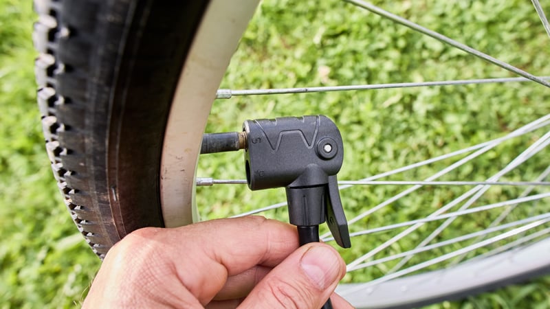 Nahaufnahme einer Hand, die eine Luftpumpendüse am Ventil eines Fahrradreifens hält. Im Hintergrund grünes Gras, die Speichen des Fahrradrades sind sichtbar.