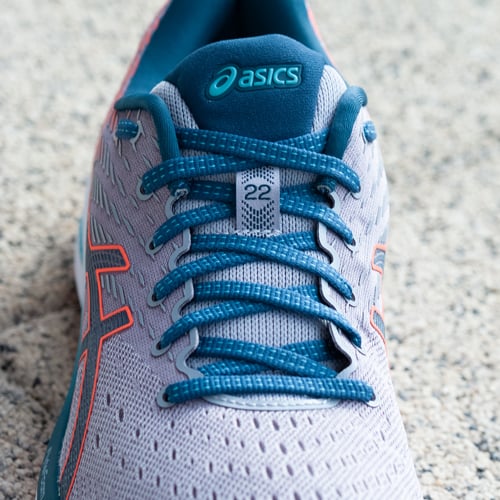 Nahaufnahme eines Asics-Laufschuhs in Hellgrau mit blauen Schnürsenkeln und blauen Akzenten. Auf der Zunge sind das Asics-Logo und die Zahl ‚22‘ zu sehen. Der Hintergrund besteht aus einer strukturierten Oberfläche, vermutlich Beton oder Kies