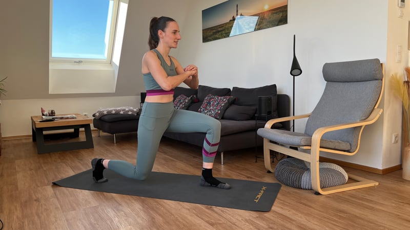 Person in Sportkleidung macht einen Ausfallschritt auf einer Yogamatte in einem Wohnzimmer mit Holzboden