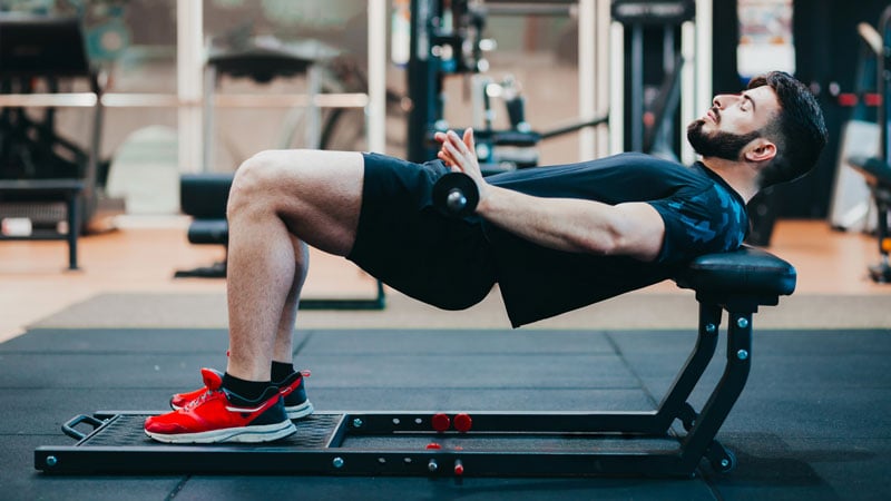 Person führt auf einer Trainingsbank im Fitnessstudio eine Hüftstoß- oder Glute-Bridge-Übung aus. Sie liegt mit gebeugten Knien und flachen Füßen auf dem Boden, hält eine Kurzhantel über der Hüfte. Im Hintergrund sind diverse Fitnessgeräte und Maschinen sichtbar