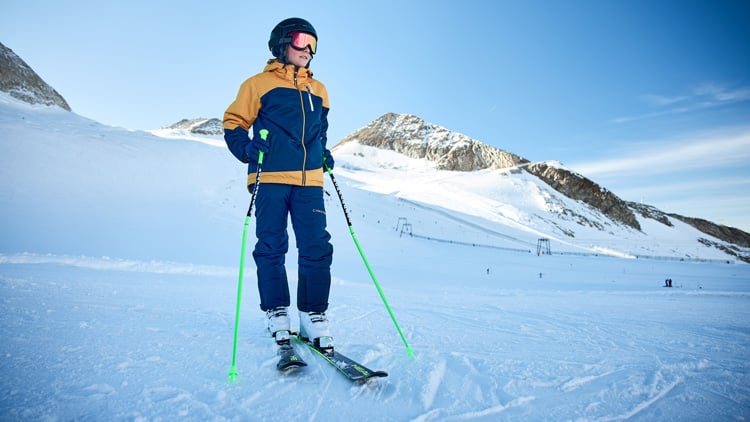 Kind steht auf Skiern in einem verschneiten Gebirgsgebiet, trägt eine blau-gelbe Jacke, blaue Hose, Skischuhe und hält grüne Skistöcke. Im Hintergrund sind schneebedeckte Berge unter klarem, blauem Himmel zu sehen