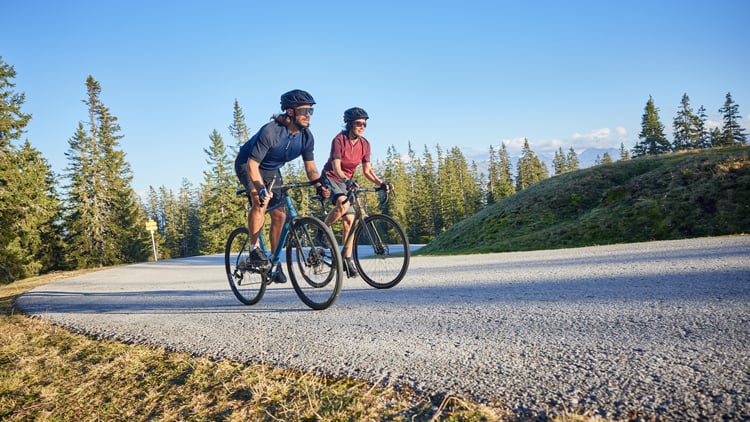 Zwei Personen auf Gravelbikes fahren auf einer Straße durch bergige Landschaft mit Bäumen und klarem Himmel; beide tragen Helme und Fahrradbekleidung.