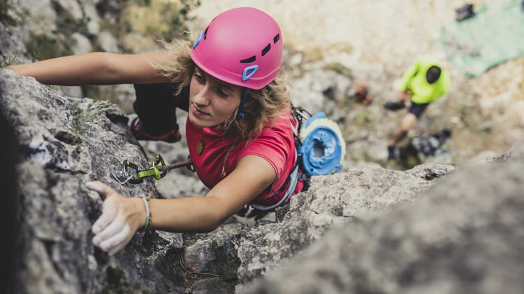 Eine Person beim Felsklettern mit pinkem Helm und rotem Shirt, gesichert mit Gurt und Karabinern. Im Hintergrund steht eine weitere Person in bunter Kleidung am Boden, vermutlich beim Sichern.