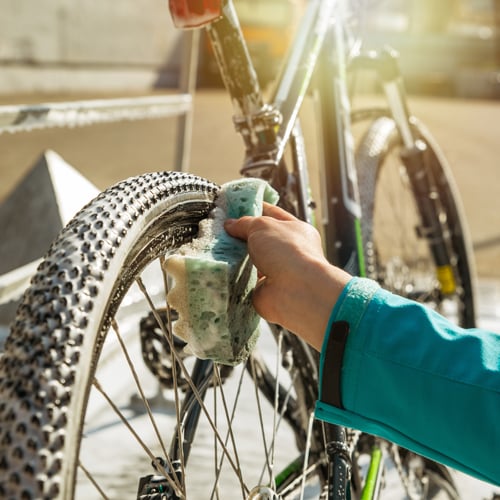 Person reinigt das Hinterrad eines Fahrrads mit einem grünen Schwamm; Wasser und Seifenschaum sind sichtbar, im Hintergrund ein sonniger Außenbereich.
