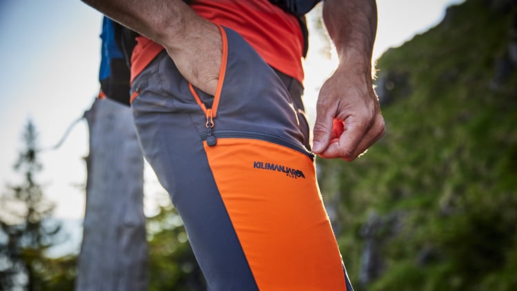 Nahaufnahme einer Person in orange-grauer Outdoorhose mit der Aufschrift KILIMANJARO. Die Person öffnet mit der rechten Hand den Reißverschluss einer Hosentasche. Im Hintergrund sind Bäume und Grünflächen zu sehen, was auf eine Wanderumgebung hinweist.