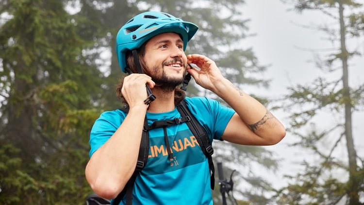 Person mit blauem Helm der Marke Kilimanjaro und blauem Shirt mit der Aufschrift ‚KILIMANJARO‘, beim Einstellen des Helms in nebliger Waldumgebung.