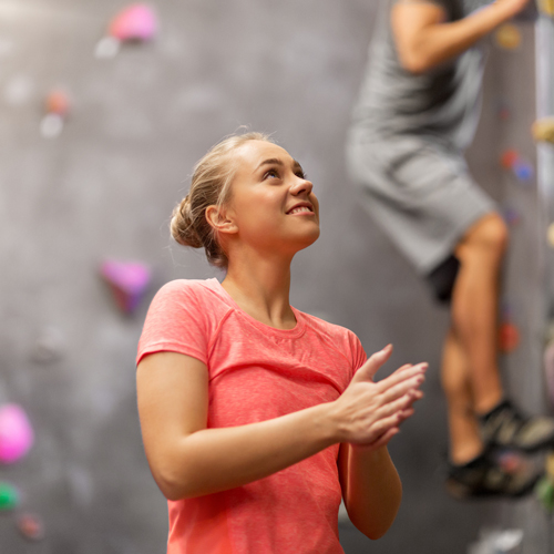Zwei Personen beim Indoor-Klettern. Im Vordergrund steht eine Person mit pinkem Shirt, die sich offenbar mit Chalk vorbereitet. Ihr Gesicht ist unscharf. Im Hintergrund klettert eine weitere Person in grauer Kleidung an einer Wand mit bunten Griffen.
