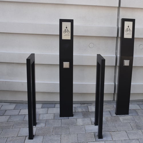 E-Bike-Ladestation mit zwei schwarzen, vertikalen Pfosten, jeweils mit einem Schild mit Fahrrad-Symbol und der Aufschrift ‚PARKING‘; davor zwei rechteckige Rahmen zum Abschließen von Fahrrädern; gepflasterter Boden und Betonwand im Hintergrund.