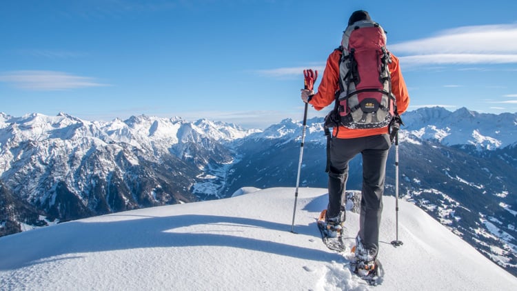 Eine Person mit Schneeschuhen und Rucksack steht auf einem verschneiten Berggipfel. Sie hält Wanderstöcke und blickt auf ein weites, schneebedecktes Gebirge unter klarem blauem Himmel