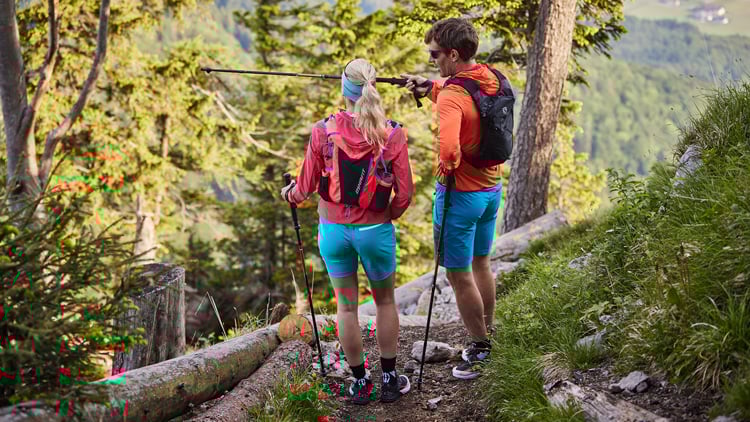 Zwei Wandernde mit Rucksäcken und Trekkingstöcken auf einem Waldpfad in den Bergen; links eine Person in pinker Jacke, rechts eine in orangefarbener Jacke, beide mit blauen Shorts und schwarzen Schuhen, im Hintergrund Bäume und Berglandschaft