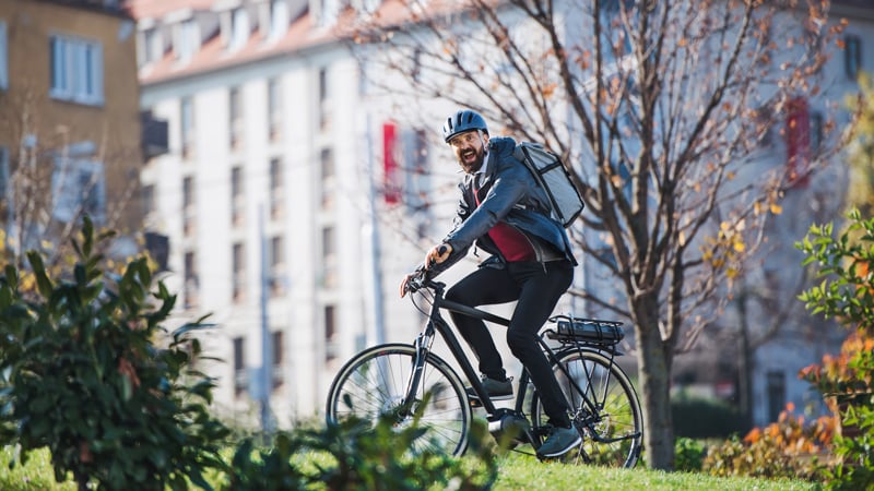 Person mit Helm, Jacke und Rucksack fährt mit dem E-Bike in einem städtischen Park bergauf. Im Hintergrund Gebäude mit vielen Fenstern und herbstlich gefärbte Bäume.