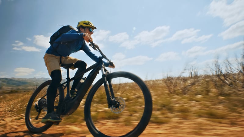 Person fährt schnell mit einem e-Mountainbike auf einem Schotterweg in einer offenen, grasbewachsenen Landschaft. Im Hintergrund sind ein klarer Himmel mit einigen Wolken und entfernte Berge zu sehen.