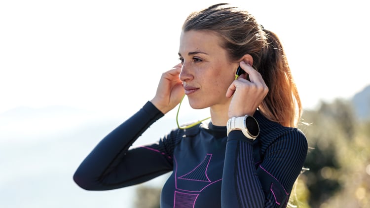 Person mit langärmligem Sportshirt mit pinken Mustern und Smartwatch am linken Handgelenk passt grüne Ohrhörer an. Im Hintergrund ist Natur zu erkennen