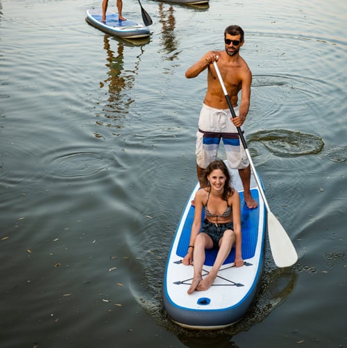 Zwei Personen befinden sich auf einem Paddleboard im Wasser – eine steht und paddelt, die andere sitzt. Im Hintergrund sind zwei weitere Personen auf einem zweiten Paddleboard zu sehen. Das Wasser ist ruhig mit leichten Wellen