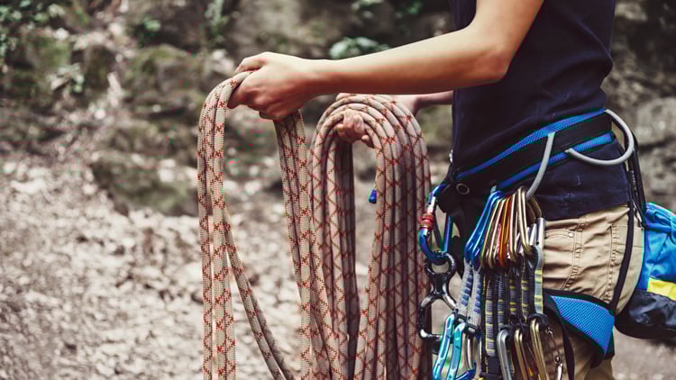 Eine Person steht im Freien mit Kletterausrüstung, hält ein Kletterseil in den Händen und trägt einen Klettergurt mit mehreren Karabinern und Expressschlingen. Im Hintergrund sind Felsen und Waldboden zu sehen.
