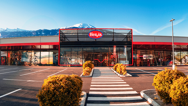 Die Außenansicht des Hervis Stores Innsbruck. Der Store hat einen großen Glaseingang mit dem Hervis-Logo, das gut sichtbar darüber angebracht ist. Vor dem Store befindet sich ein Parkplatz mit mehreren Büschen und einem Fußgängerüberweg, der zum Eingang führt. Im Hintergrund sind unter einem klaren blauen Himmel Berge zu sehen.