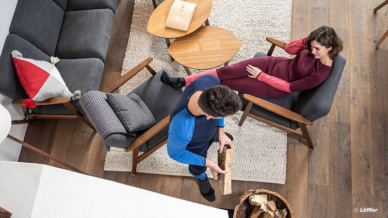 Wohnzimmer mit zwei Personen – eine sitzt auf einem Stuhl, die andere steht und hält ein Stück Feuerholz. Der Raum hat einen Holzboden, ein graues Sofa mit einem rot-weißen Kissen, zwei runde Holztische und einen Korb mit Feuerholz. Foto: Löffler.