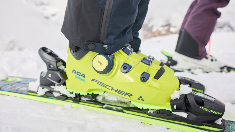 Skischuhe richtig lagern Reihe von Skiern und Snowboards, die auf einem verschneiten Berghang aufrecht in Holzständern stehen. Im Hintergrund sind schneebedeckte Berge unter klarem, blauem Himmel zu sehen