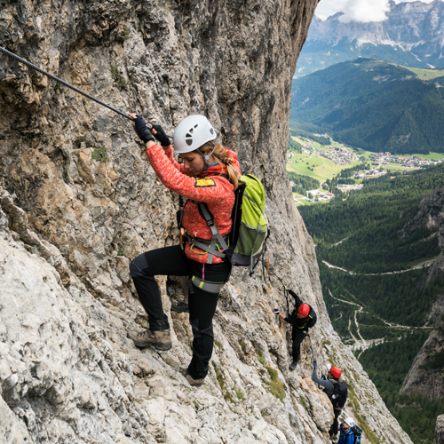 Drei Personen beim Klettern an einer steilen Felswand. Sie tragen Helme, Gurte und Rucksäcke und sichern sich mit einem Fixseil. Im Hintergrund ist eine malerische Landschaft mit grünen Tälern und Bergen unter einem teils bewölkten Himmel zu sehen.