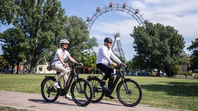 Zwei Personen fahren mit e-Bikes der Marke KTM durch einen Park mit grünem Rasen und Bäumen; im Hintergrund steht ein großes Riesenrad. Beide tragen Helme und Freizeitkleidung. Copyright: KTM