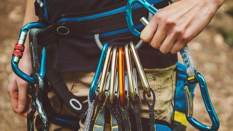 Ein Kletterer hält eine Gruppe bunter Karabiner, die an einem Klettergurt befestigt sind. Der Gurt ist mit mehreren Sicherungsgeräten ausgestattet. Im Hintergrund ist eine unscharfe, natürliche Umgebung zu erkennen, vermutlich ein Wald oder Felsgebiet.